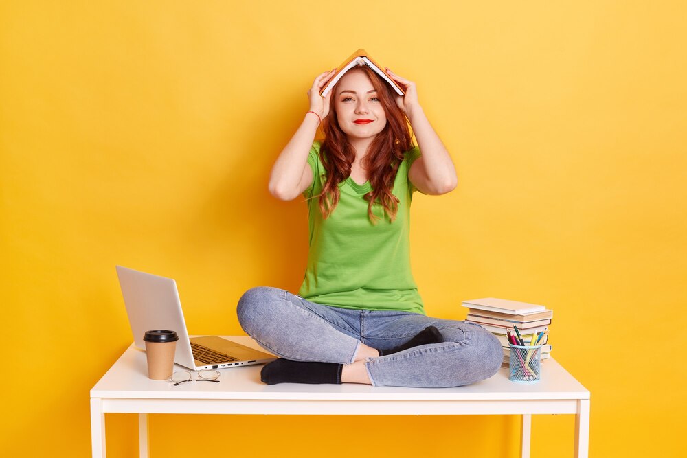 smiling-young-european-female-sitting-table-with-crossed-legs-make-roof-with-book-her-head-looks-camera-wearing-casually-surrounding-with-books-laptop-coffee_176532-13955.jpg